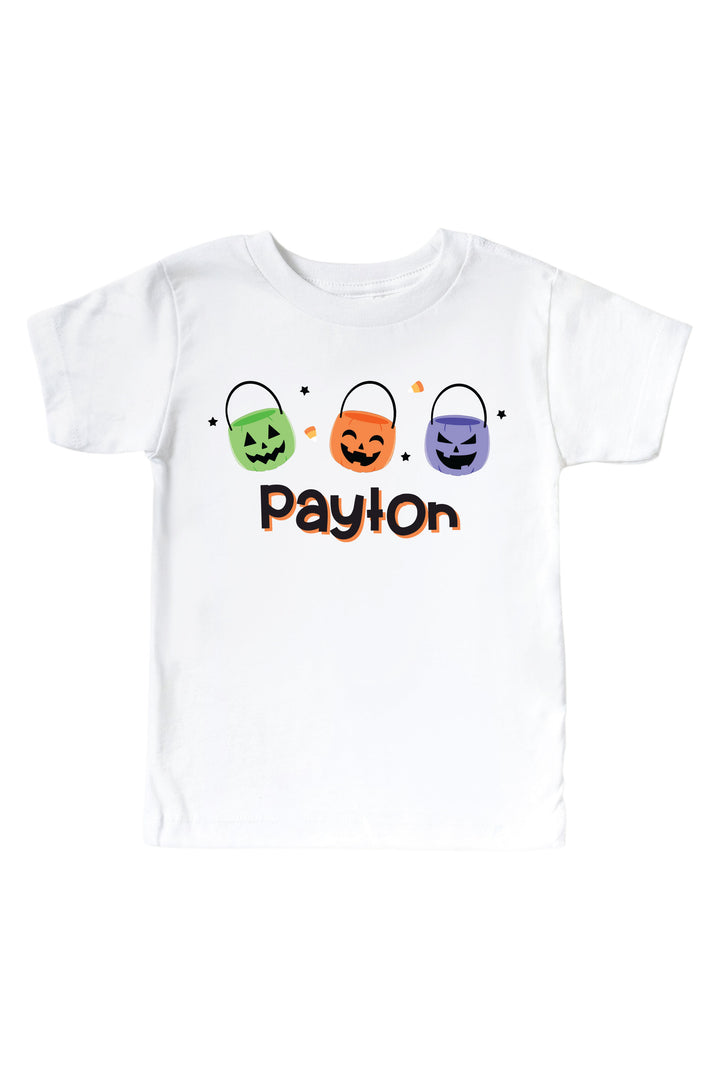 White kids' t-shirt and matching bodysuit personalized with names in orange script below three colorful jack-o-lantern candy bucket icons