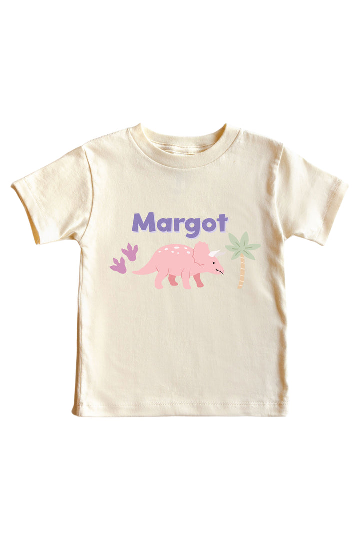 Natural cream kids tee with personalized name in purple lettering and a pink triceratops dinosaur graphic with pastel plant accents
