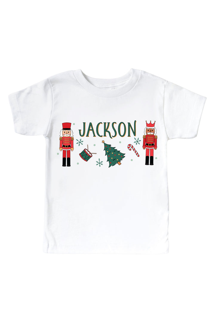 Caden Lane's white baby onesie personalized with the name "Jackson," featuring red toy soldier nutcrackers, a Christmas tree, drum, and candy cane, laid flat.