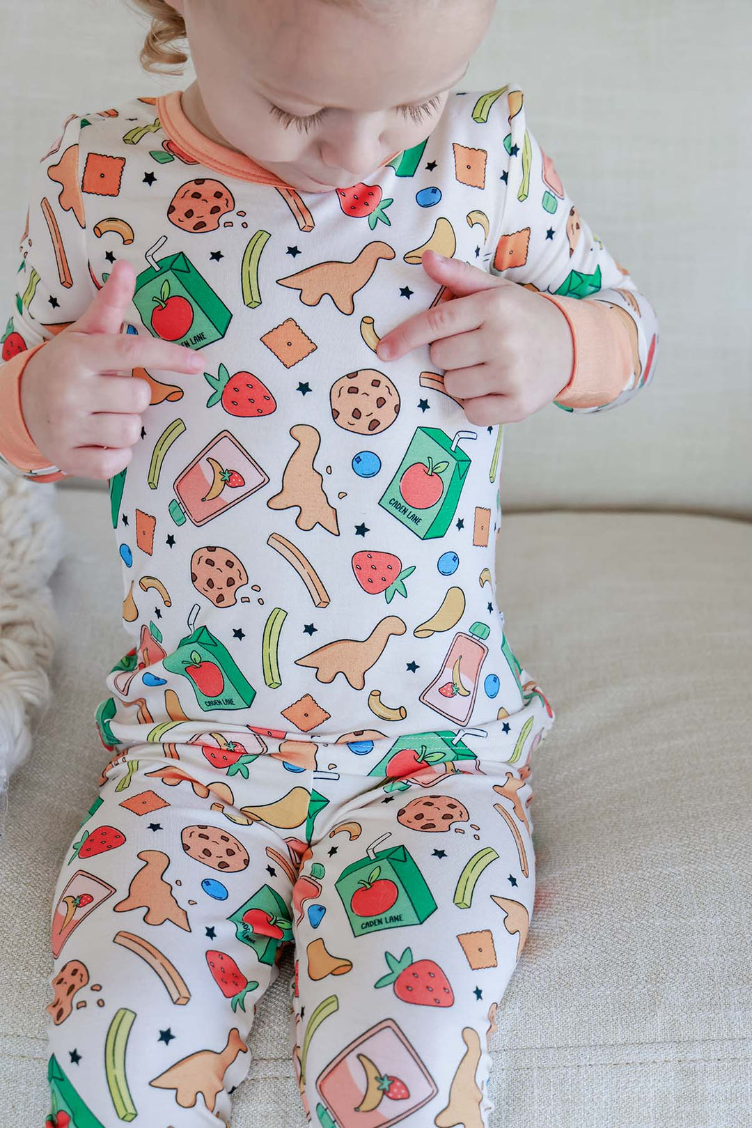 Toddler sitting and wearing the Snack Attack Two Piece Bamboo Pajama Set, showing the long-sleeve top with playful snack print featuring cookies, strawberries, dinosaur crackers, juice cartons, and bananas with peach orange trim