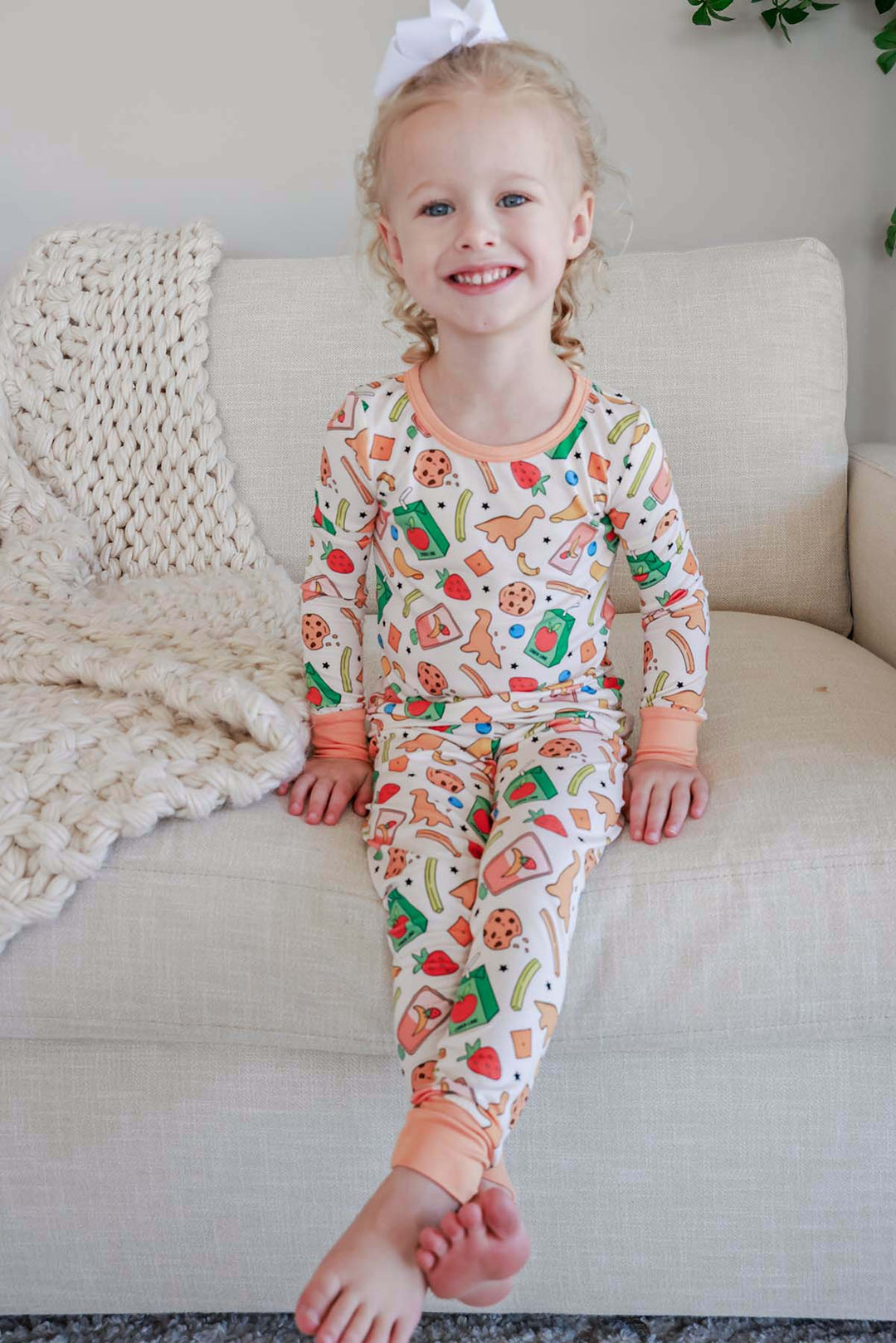 Smiling young girl with a white bow sitting on a cream couch wearing the Snack Attack Two Piece Bamboo Pajama Set, showing both the long-sleeve top and matching pants with peach orange trim and the colorful snack print