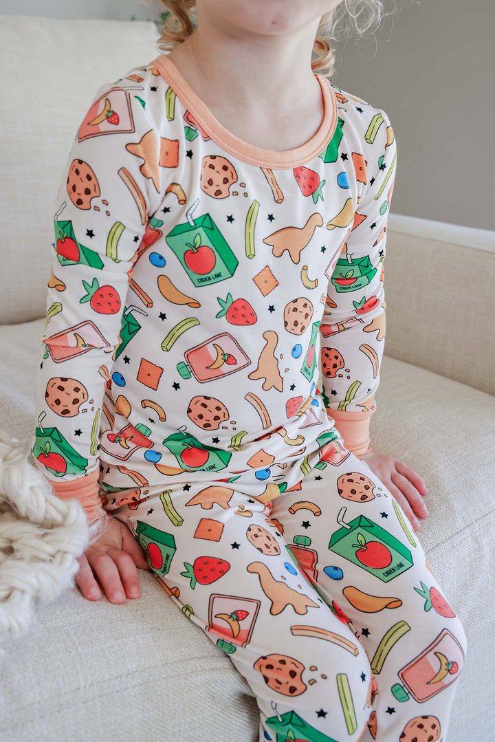 Young girl sitting on a couch wearing the full Snack Attack Two Piece Bamboo Pajama Set, displaying both the matching long-sleeve top and pants with peach orange crew neck trim and the colorful snack print