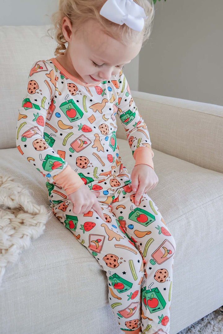 Sibling lifestyle photo of a smiling big sister with a white bow holding a sleeping newborn with white bow headband, both in coordinating Snack Attack bamboo pajamas from Caden Lane