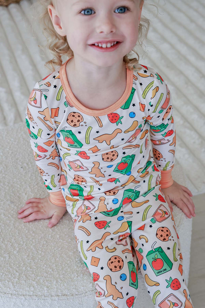 Big sister smiling while cradling a sleeping newborn, both wearing coordinating Snack Attack print bamboo pajamas - big sister in the Two Piece Set and newborn in the Ruffle Bamboo Zipper Footie