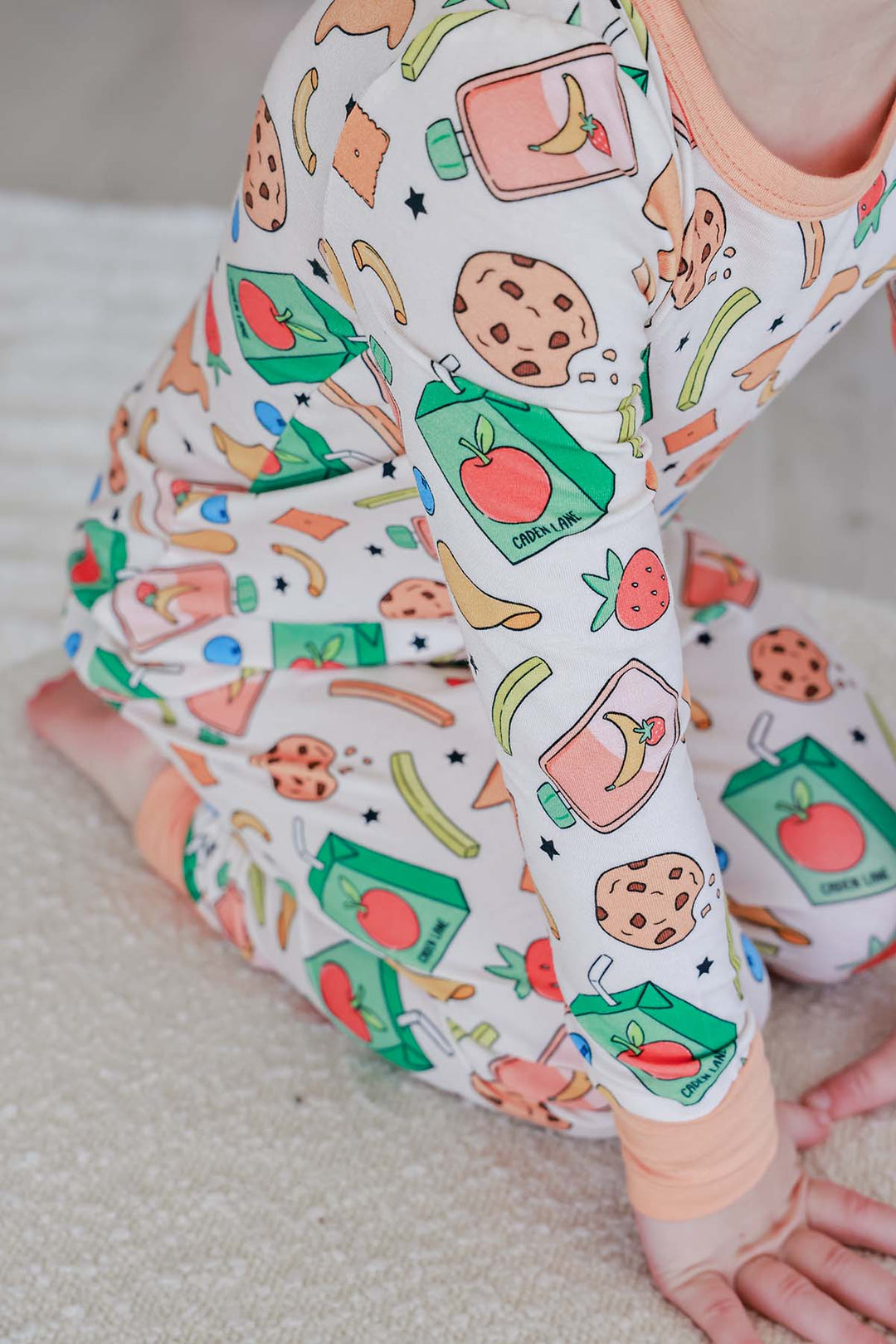 Two sisters in coordinating Snack Attack bamboo sleepwear - older sister holding sleeping baby, both in the playful snack-themed print, showing Caden Lane's matching sibling pajama options