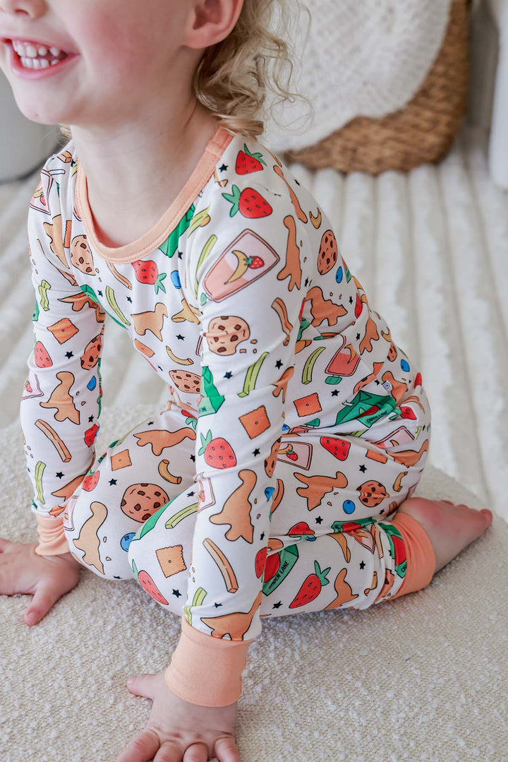 Lifestyle sibling photo showing big sister with white bow holding a sleeping newborn, both wearing the Snack Attack print bamboo collection from Caden Lane, highlighting the coordinating sleepwear collection