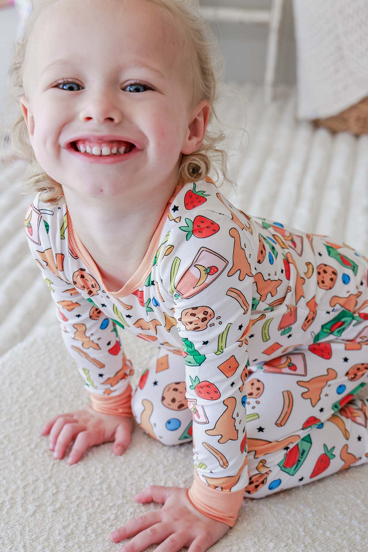 Smiling young girl leaning forward wearing the Snack Attack Two Piece Bamboo Pajama Set, featuring a close view of the long-sleeve top's snack print with cookies, strawberries, dinosaur crackers, and banana