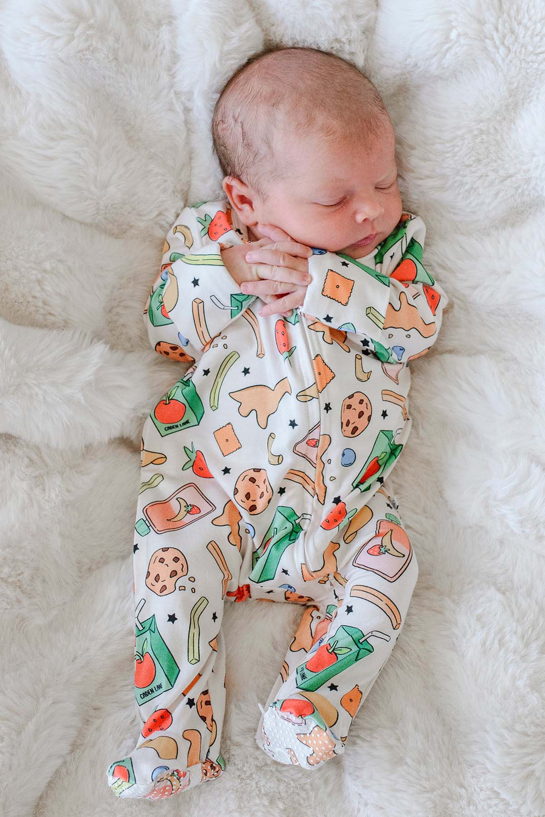 Sleeping newborn lying on a white fluffy blanket wearing the Snack Attack Bamboo Zipper Footie, showing the snack-themed print with cookies, dinosaur crackers, juice cartons, bananas, and strawberries