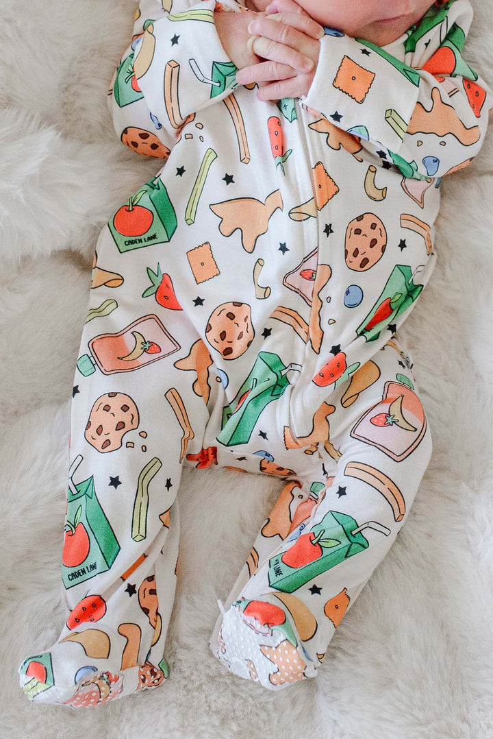 Overhead view of a newborn's torso to feet lying on a fluffy cream blanket in the Snack Attack Bamboo Zipper Footie, with the snack print clearly displaying dinosaur crackers, juice cartons, cookies, and banana