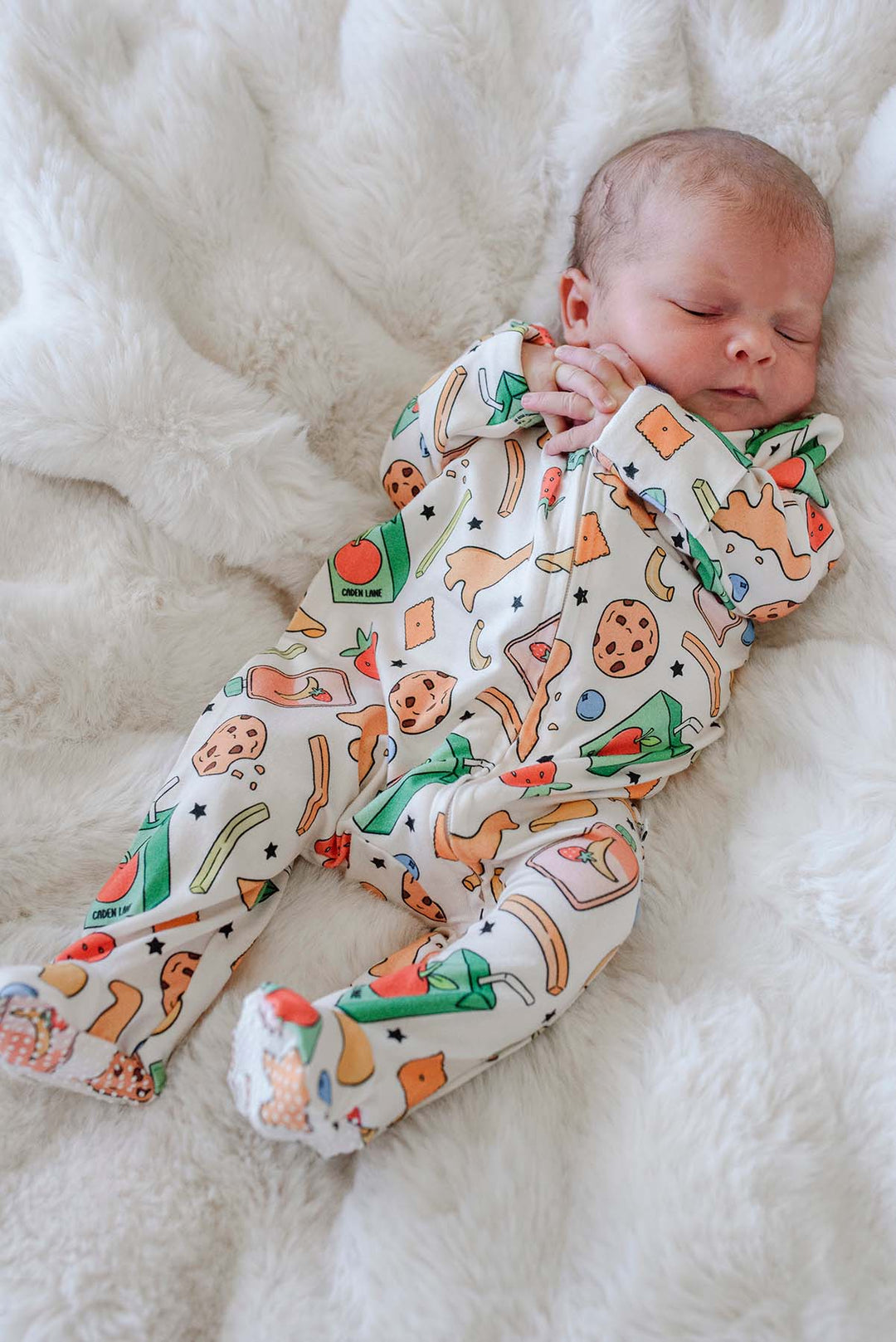 Full-body view of a sleeping newborn on a white fluffy blanket wearing the Snack Attack Bamboo Zipper Footie, showing the snack print with cookies, dinosaur crackers, juice cartons, banana, and watermelon from head to feet