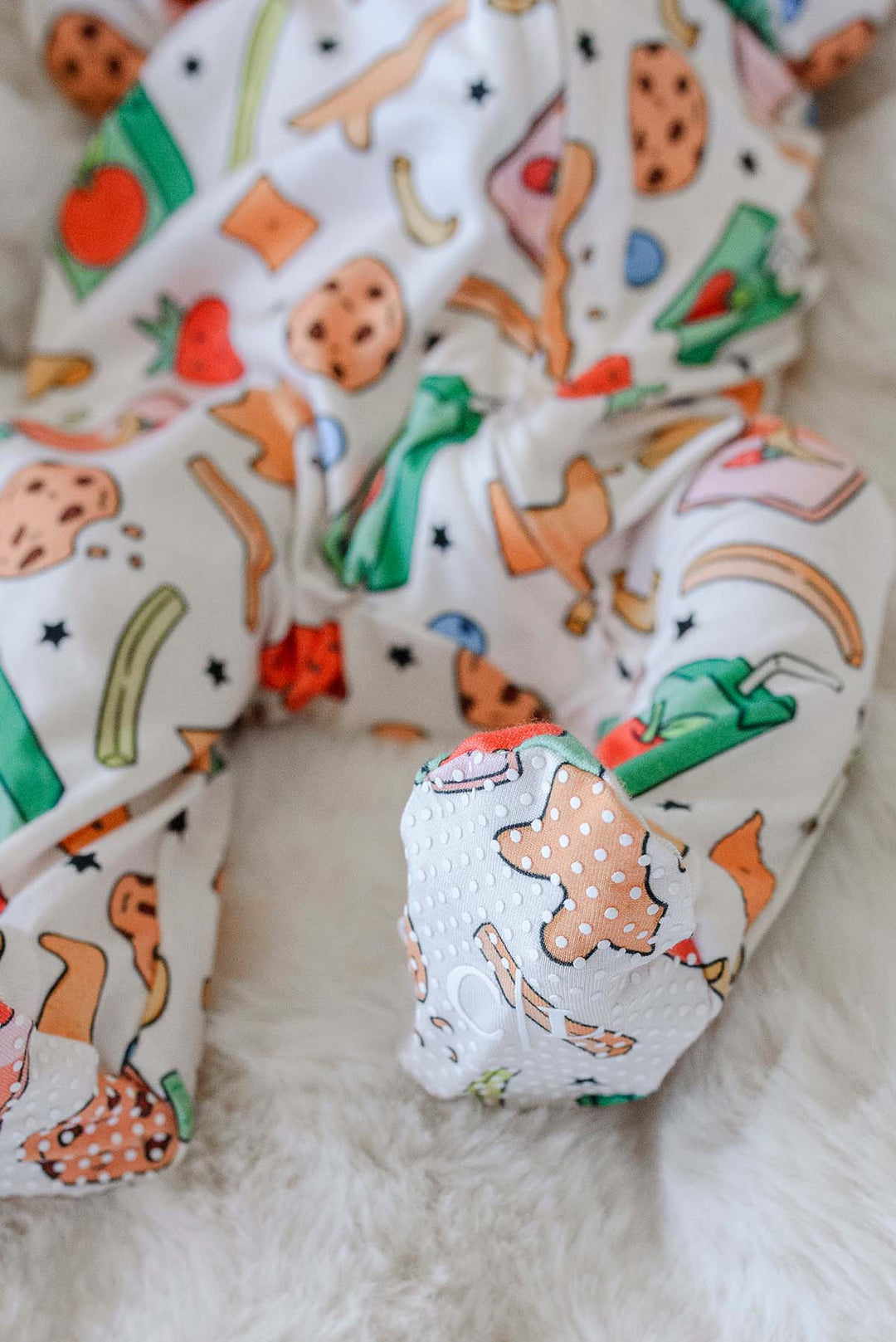 Close-up of a newborn's feet and lower legs in the Snack Attack Bamboo Zipper Footie, showing the footed design with grip-print sole and the snack print with dinosaur crackers, cookies, and strawberries