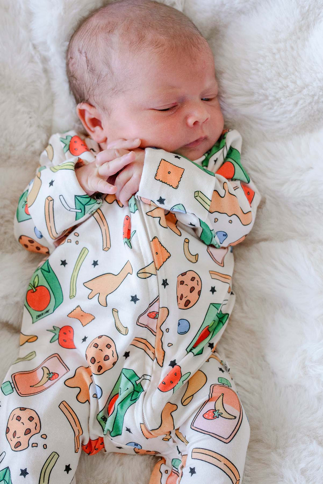Close view of a sleeping newborn's head and upper body on a white fluffy blanket wearing the Snack Attack Bamboo Zipper Footie, showing the vibrant snack print with apple juice carton, dinosaur crackers, cookies, and strawberries