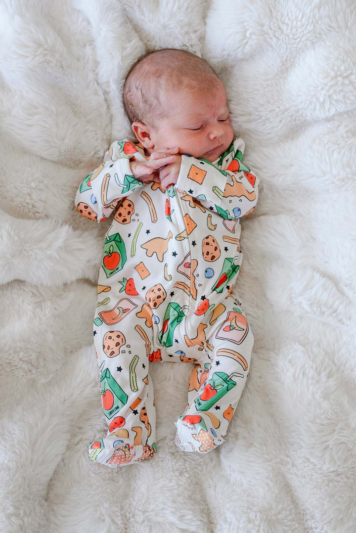 Sleeping newborn with hand near face, lying on a white fluffy blanket wearing the Snack Attack Bamboo Zipper Footie, showing the full snack-themed bamboo print from head to toe