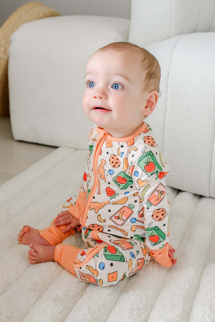 Baby seated and looking down at a chocolate chip cookie wearing the Snack Attack Convertible Bamboo Zip Romper in footless mode with the convertible cuffs open, displaying the vibrant snack-themed print