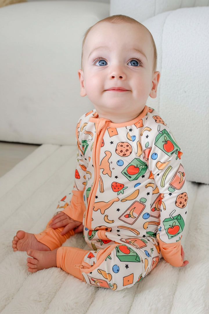Baby with blue eyes sitting upright wearing the Snack Attack Convertible Bamboo Zip Romper, showing the playful cookie, strawberry, banana, juice carton, and dinosaur cracker print with peach orange zip and cuffs