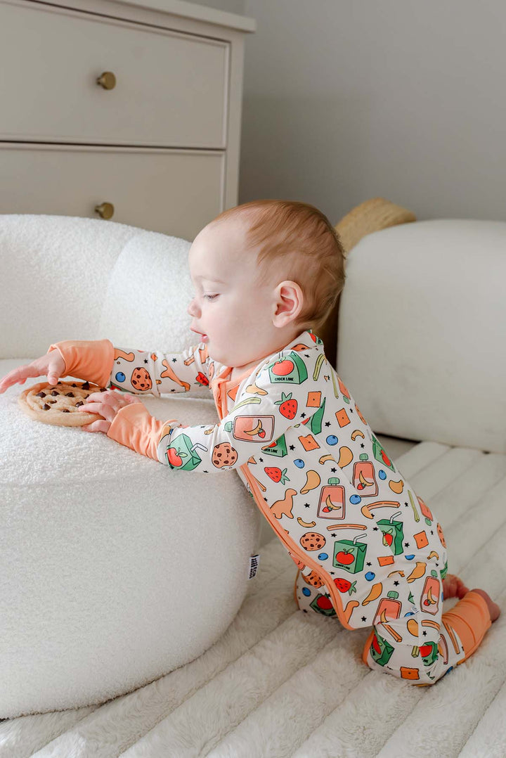 Baby reaching forward and playing with a wooden teether ring while wearing the Snack Attack Convertible Bamboo Zip Romper, displaying the colorful snack print with orange peach zip detail