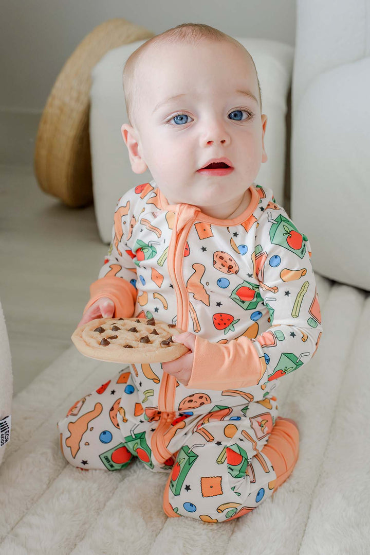 Baby with blue eyes sitting and holding a large chocolate chip cookie while wearing the Snack Attack Convertible Bamboo Zip Romper with peach orange footed cuffs and snack-themed print