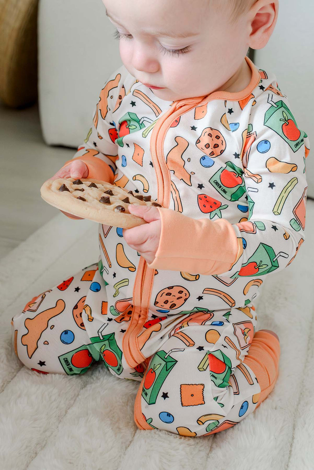 Baby seated and examining a chocolate chip cookie while wearing the Snack Attack Convertible Bamboo Zip Romper with peach orange open-toe convertible cuffs and the fun snack-themed bamboo print