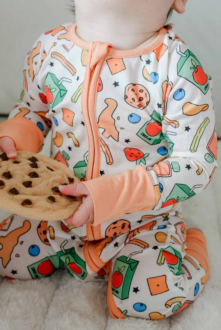 Baby holding a real chocolate chip cookie while wearing the Snack Attack Convertible Bamboo Zip Romper, showcasing the fun snack-themed print with cookies, dinosaur crackers, juice cartons, and strawberries