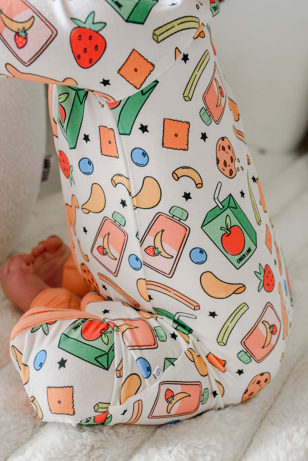 Close-up back view of a baby wearing the Snack Attack Convertible Bamboo Zip Romper, showing the detailed snack print with strawberries, crackers, bananas, and juice cartons, and the peach orange footed cuffs