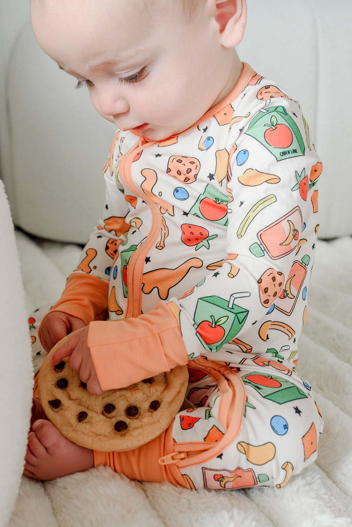 Baby looking down and holding a chocolate chip cookie while wearing the Snack Attack Convertible Bamboo Zip Romper with open-toe cuffs folded back, showing the snack print and the versatile convertible feature