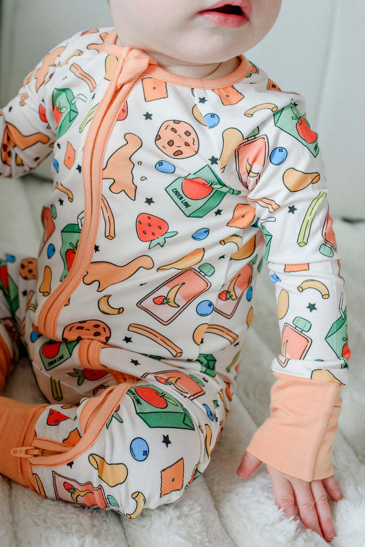Side view of baby seated with a chocolate chip cookie while wearing the Snack Attack Convertible Bamboo Zip Romper with open convertible cuffs, showing the cheerful snack print from the side