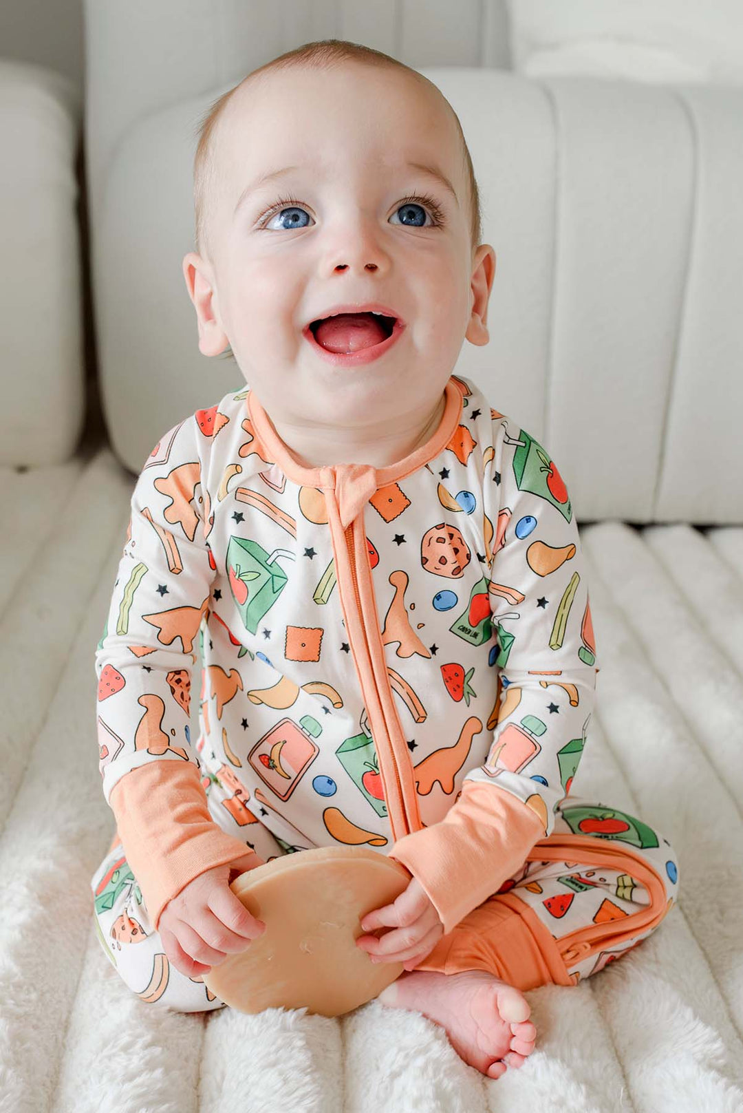 Smiling baby with blue eyes sitting upright wearing the Snack Attack Convertible Bamboo Zip Romper with peach orange zip and footed cuffs, featuring the playful snack print