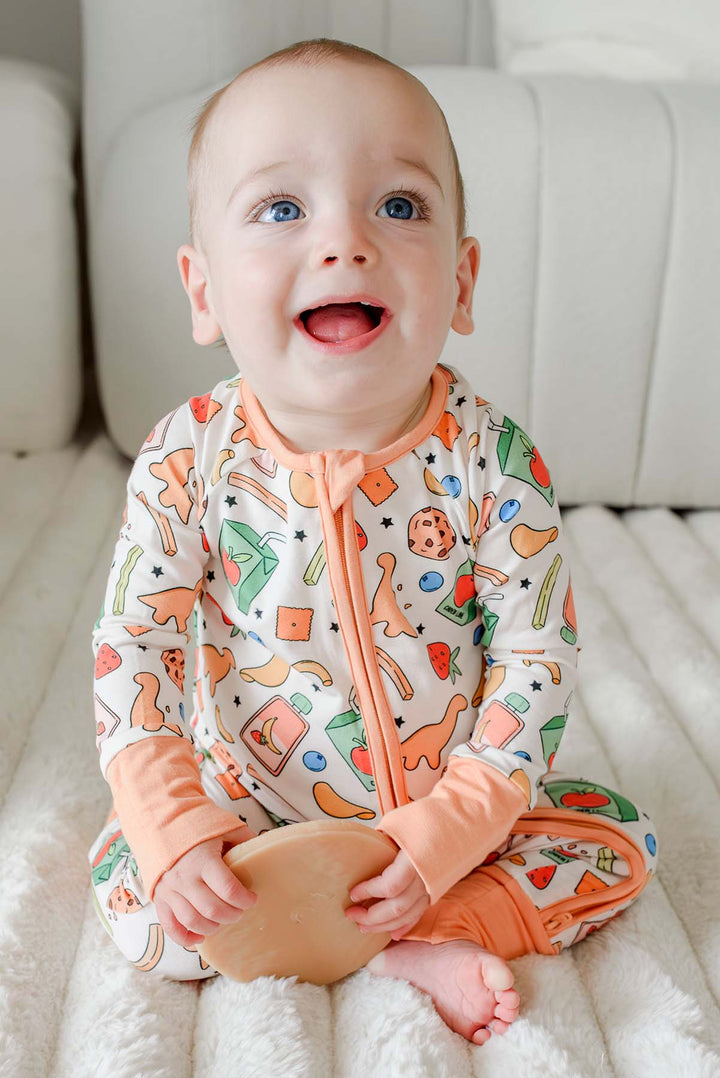 Smiling baby with blue eyes sitting upright wearing the Snack Attack Convertible Bamboo Zip Romper with peach orange zip and footed cuffs, featuring the playful snack print