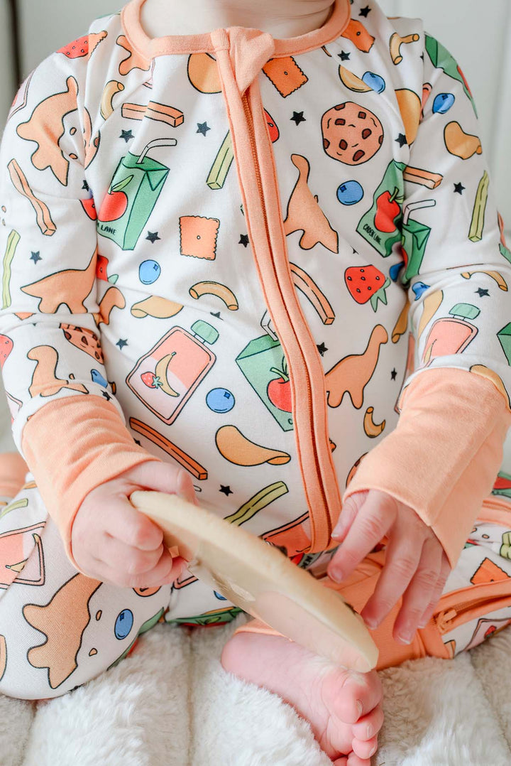 Close-up of baby wearing the Snack Attack Convertible Bamboo Zip Romper, showing the detailed snack-themed print with dinosaur crackers, cookies, strawberries, and juice cartons, and the convertible peach orange cuffs