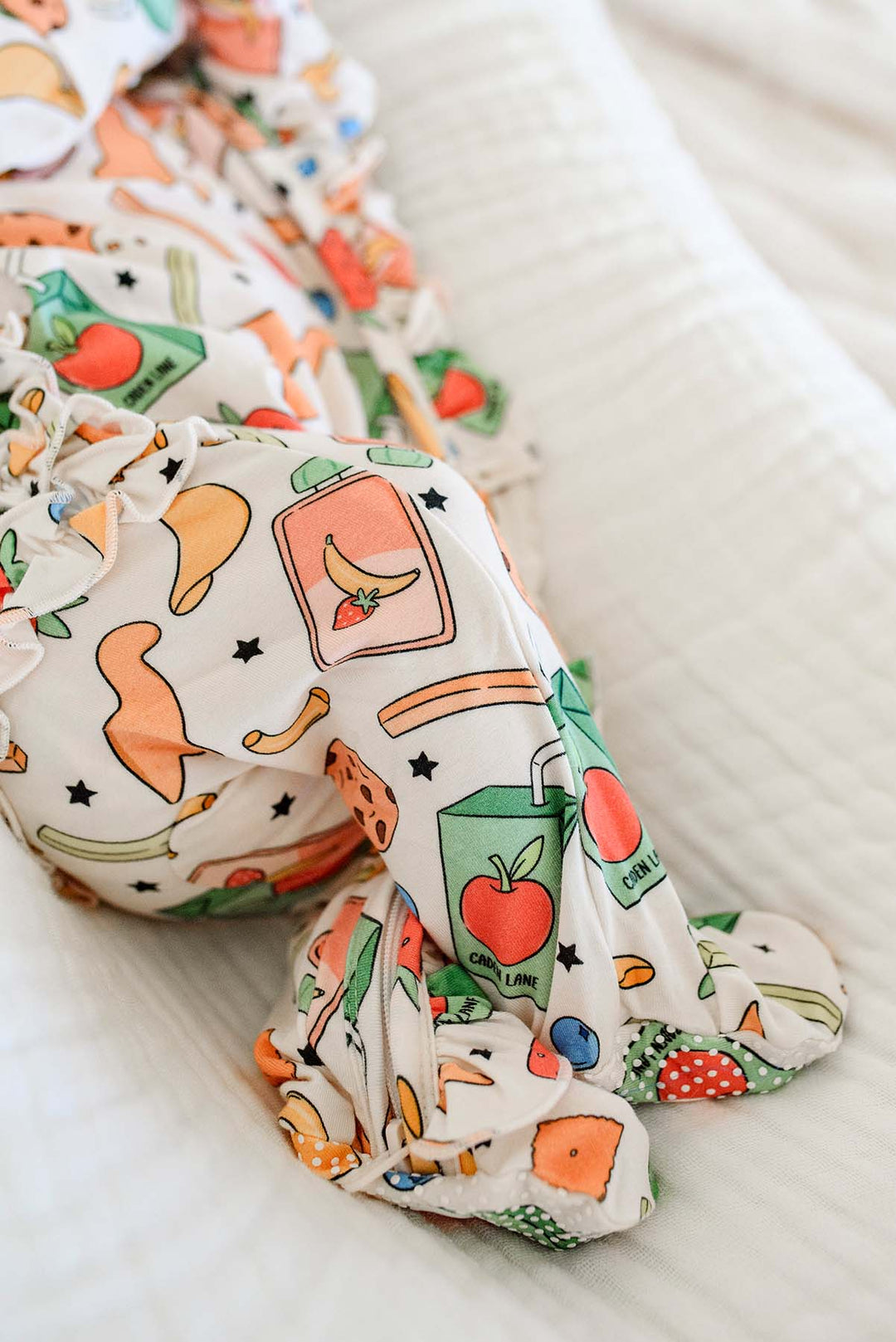 Newborn lying on back on a white textured blanket with a large white bow headband, wearing the Snack Attack Ruffle Bamboo Zipper Footie, displaying the colorful snack print bamboo sleepwear