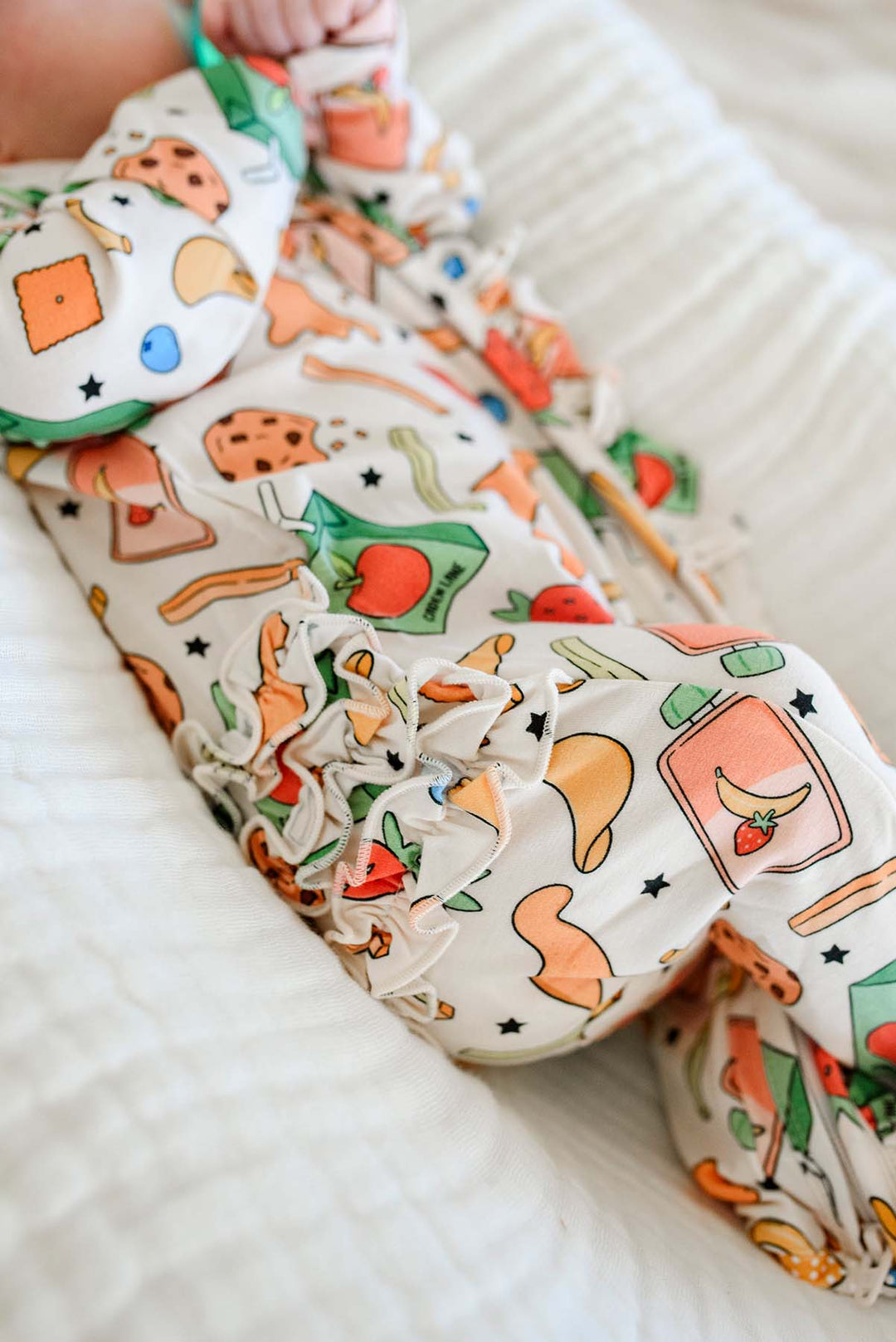 Close-up of newborn's feet and legs in the Snack Attack Ruffle Bamboo Zipper Footie, showing the ruffle bum detail and a clear view of the playful snack print including juice cartons, bananas, mushrooms, and blueberries