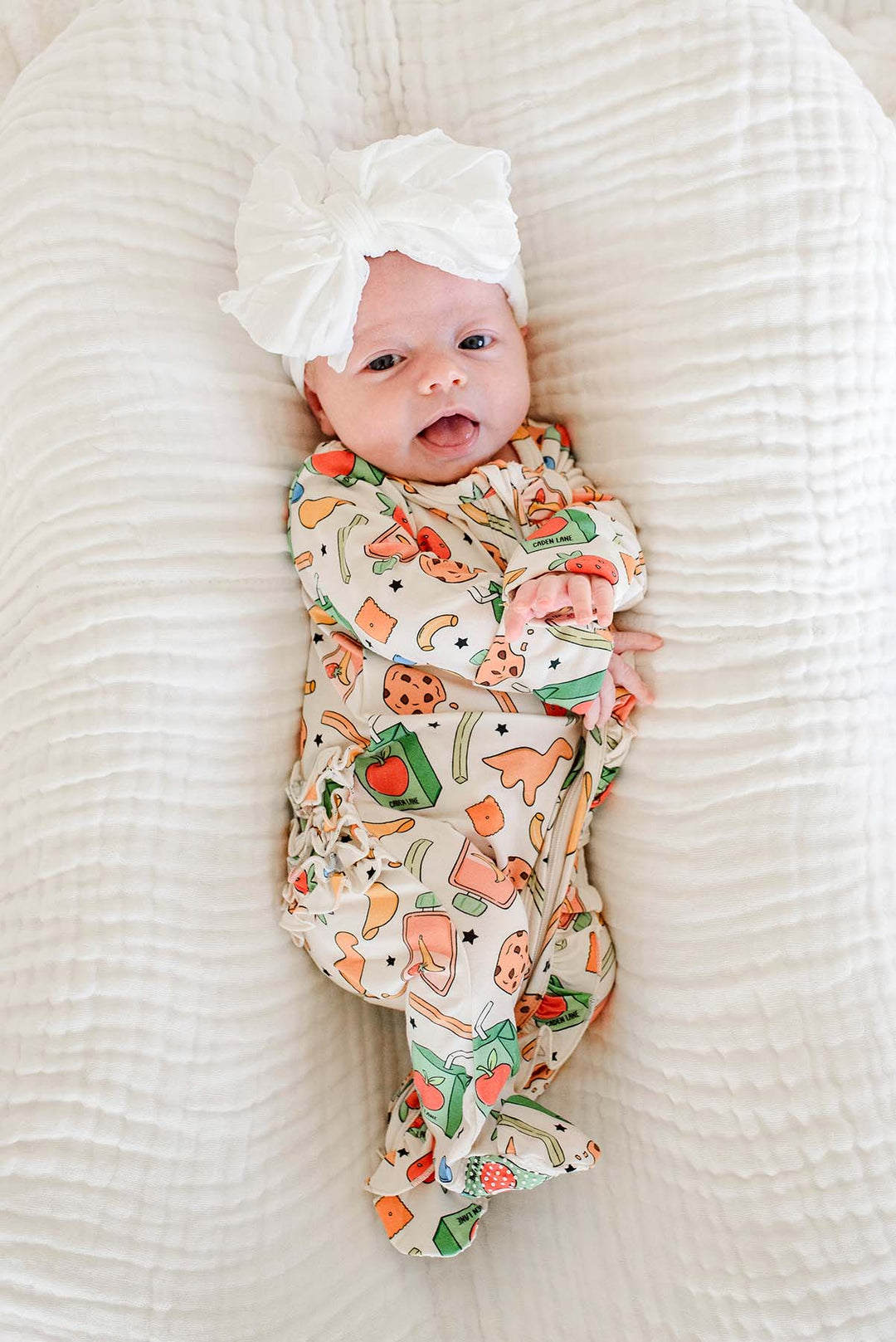 Newborn lying on back on a white textured blanket with a white bow headband, wearing the Snack Attack Ruffle Bamboo Zipper Footie, showing the playful snack print with dinosaur crackers and juice cartons