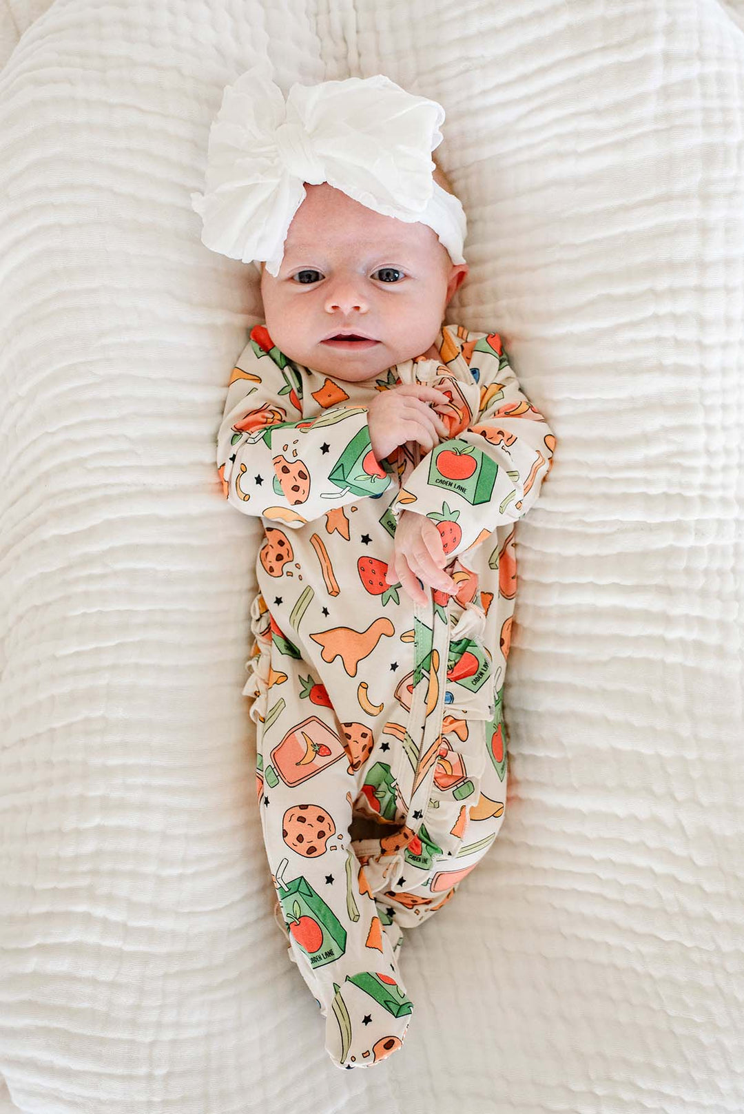 Alert newborn lying on back on a white textured blanket wearing a white bow headband and the Snack Attack Ruffle Bamboo Zipper Footie, showing the full-body snack-themed print from head to feet