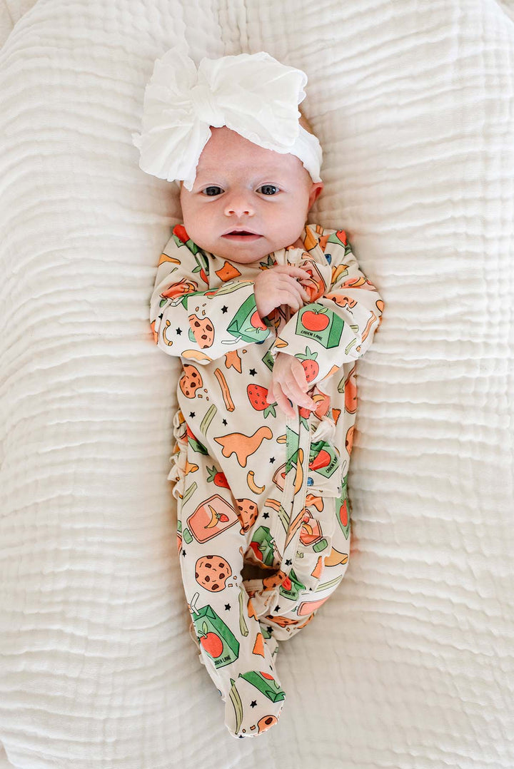 Alert newborn lying on back on a white textured blanket wearing a white bow headband and the Snack Attack Ruffle Bamboo Zipper Footie, showing the full-body snack-themed print from head to feet