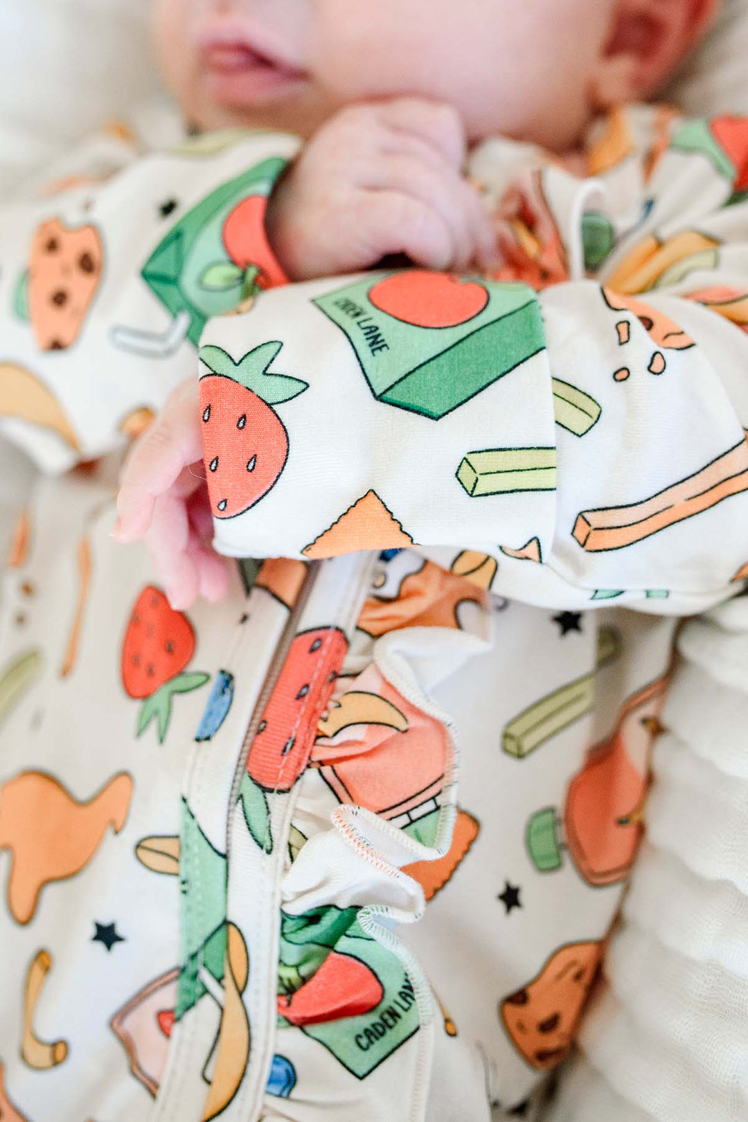 Overhead close-up of a newborn's feet and legs in the Snack Attack Ruffle Bamboo Zipper Footie, displaying the ruffle detailing and the vibrant snack-themed bamboo print in detail