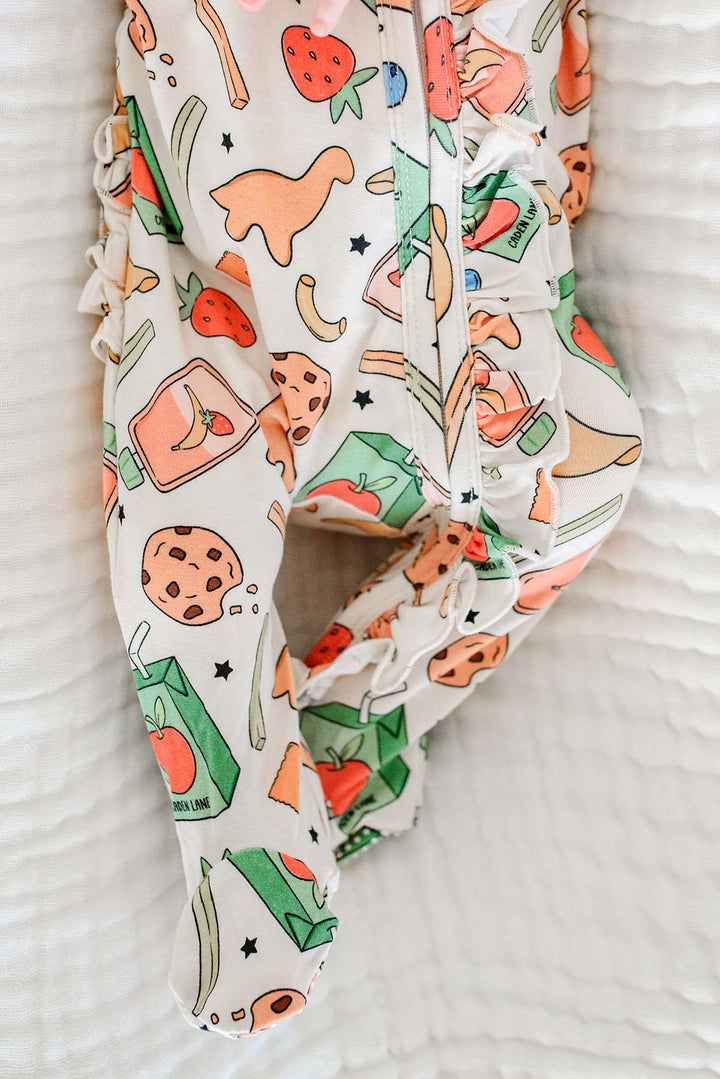 Close-up detail of newborn's legs in the Snack Attack Ruffle Bamboo Zipper Footie on a white textured blanket, highlighting the delicate ruffle trim and vibrant snack print with strawberries, dinosaur crackers, juice cartons, and cookies
