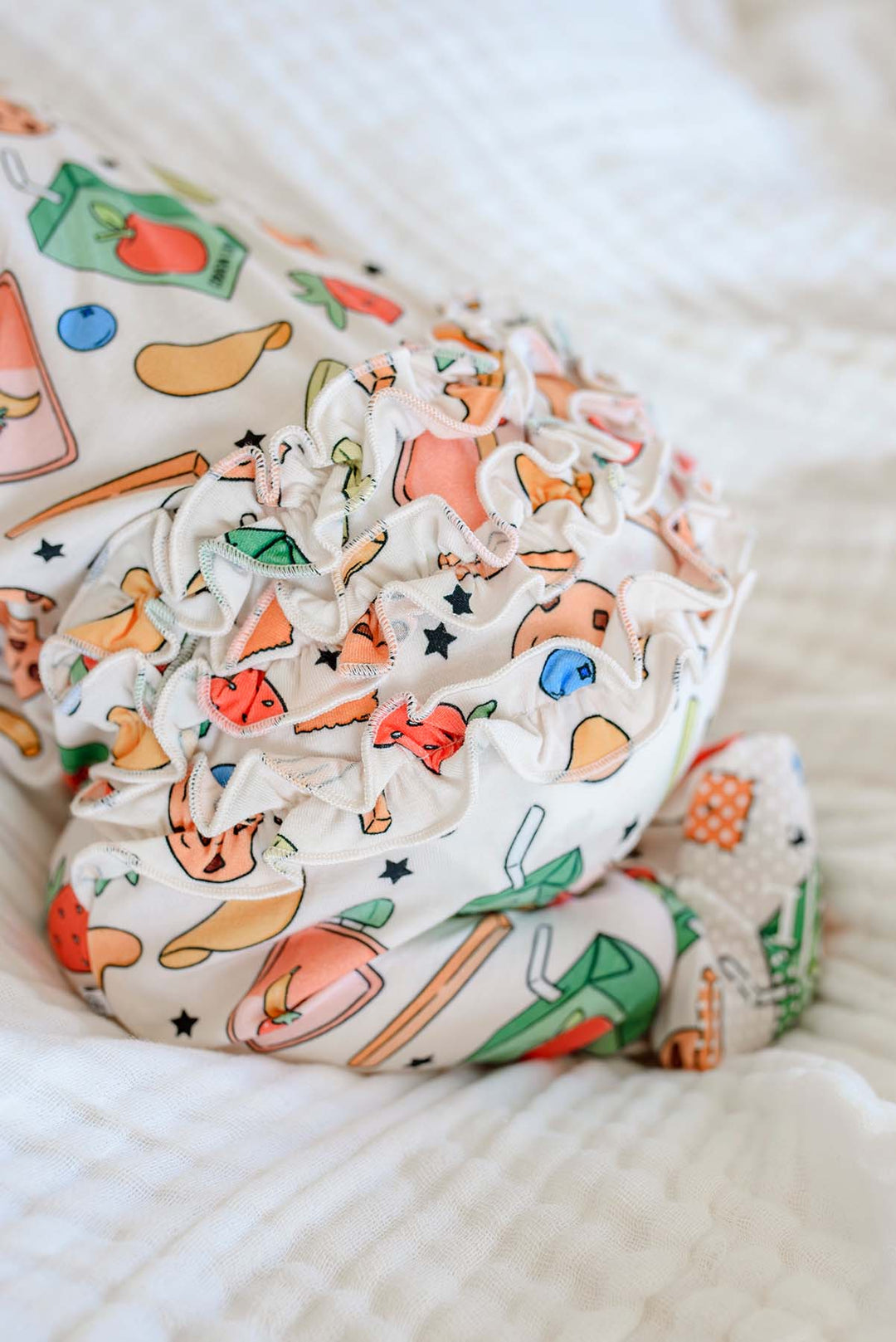 Close-up of newborn feet and legs wearing the Snack Attack Ruffle Bamboo Zipper Footie on a white blanket, featuring the ruffle bum and snack print with apple juice carton, banana, blueberries, and playful snack icons