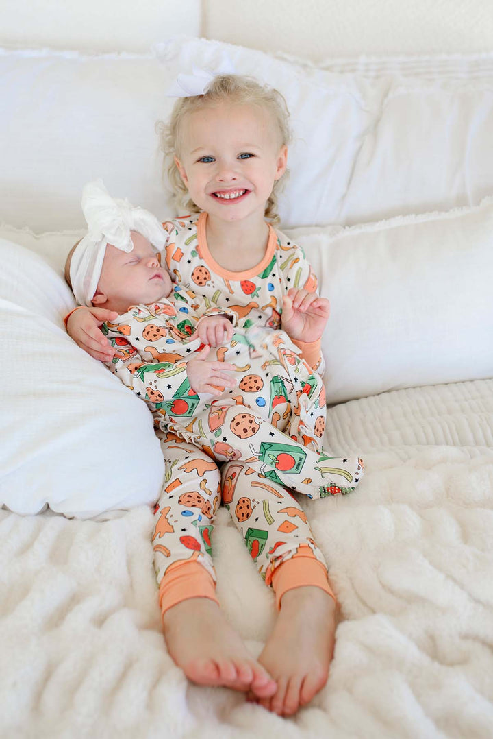 Big sister with white bow sitting on a white bed cradling a sleeping newborn, big sister wearing the Snack Attack Two Piece Bamboo Pajama Set and newborn in the Snack Attack Ruffle Footie with white bow headband