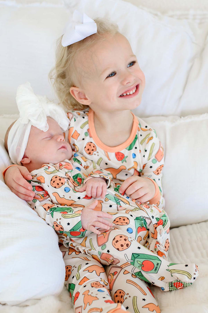 Sibling photo of big sister holding a sleeping newborn with a white bow headband, both wearing coordinating Snack Attack print bamboo sleepwear - newborn in the Bamboo Zipper Footie and big sister in the Two Piece Pajama Set