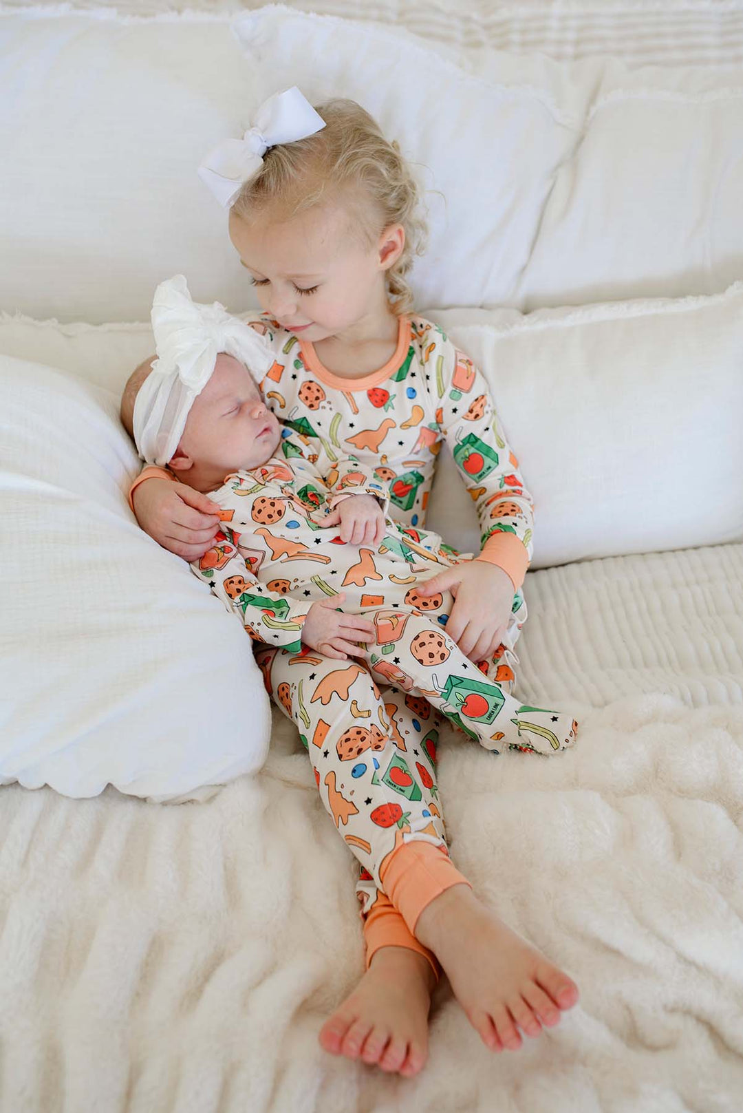 Sibling photo with big sister cradling a sleeping newborn, both in Snack Attack print bamboo sleepwear - the Two Piece Bamboo Pajama Set and Ruffle Bamboo Zipper Footie from Caden Lane