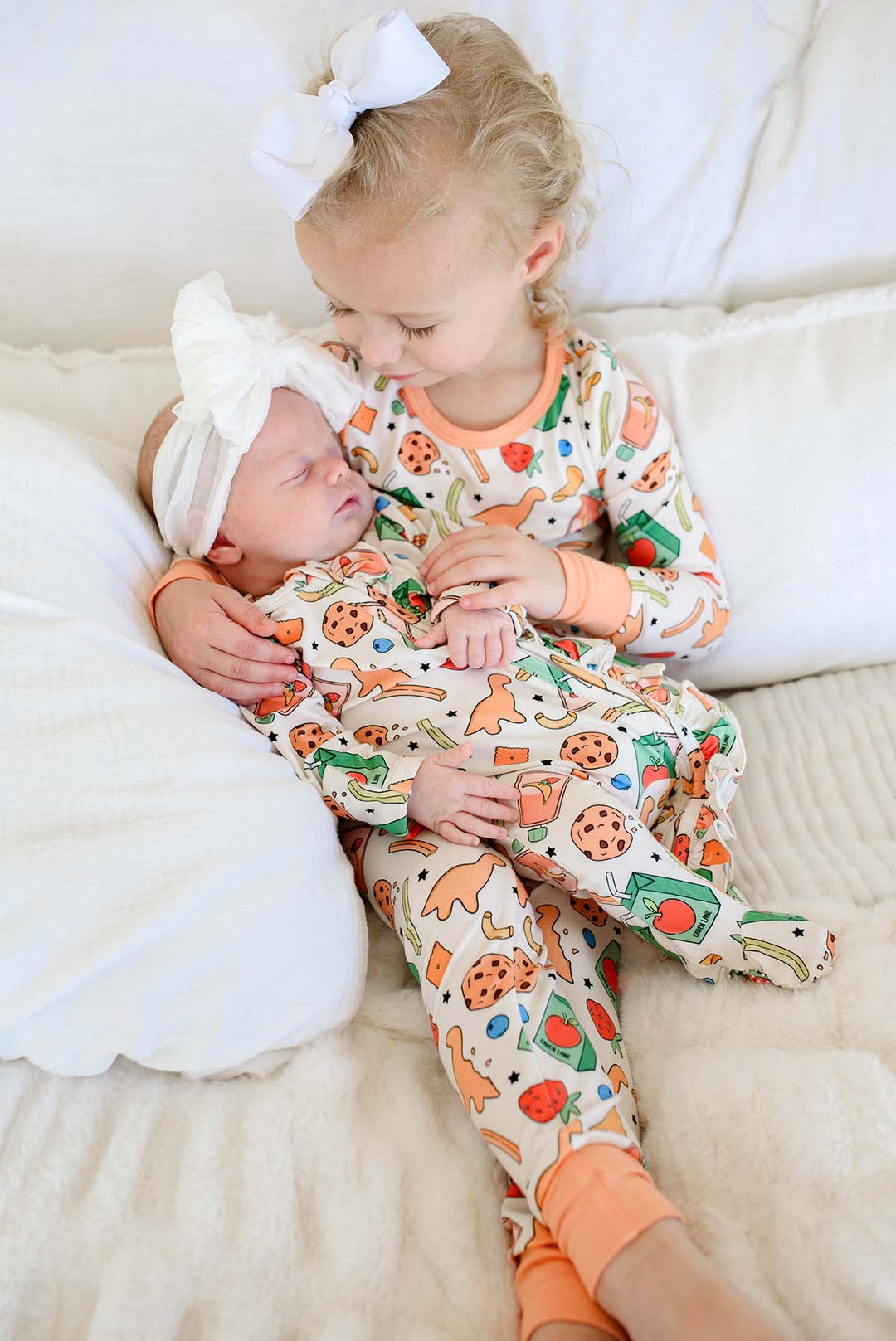 Tender sibling moment with big sister looking down lovingly at a sleeping newborn in a white bow headband, both wearing coordinating Snack Attack print bamboo sleepwear from Caden Lane