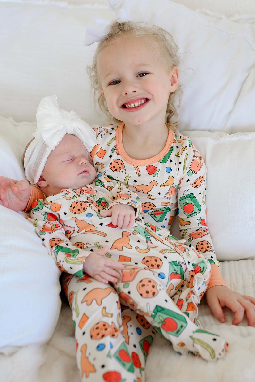 Smiling big sister holding a sleeping newborn wearing a white bow headband, big sister in the Snack Attack Two Piece Bamboo Pajama Set and newborn in the Snack Attack Ruffle Bamboo Zipper Footie, coordinating sibling outfits