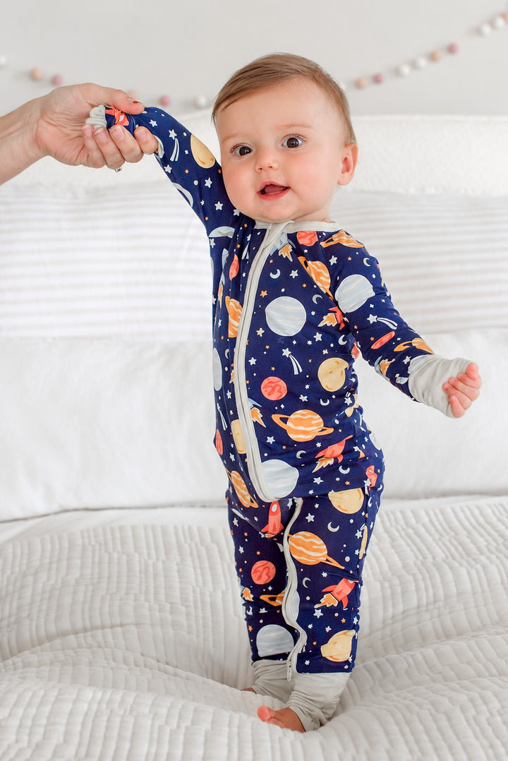 Side view of a baby standing with one hand held by an adult, wearing a Caden Lane navy bamboo zip romper in a Stellar Snuggles print featuring planets and rockets.