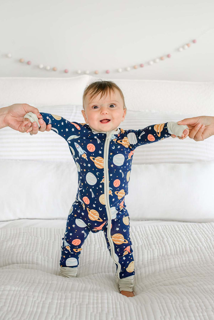 Smiling baby standing on a bed with arms outstretched and both hands held by adults, wearing a Caden Lane navy bamboo zip romper in a Stellar Snuggles print featuring planets.