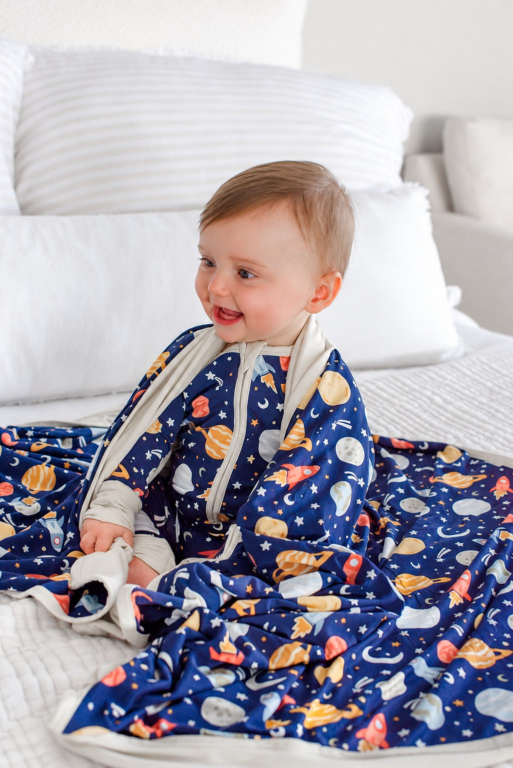 Smiling baby with a Caden Lane navy knit blanket draped over the shoulders, in a Stellar Snuggles print featuring Saturn, moons, and rockets, paired with matching pajamas.