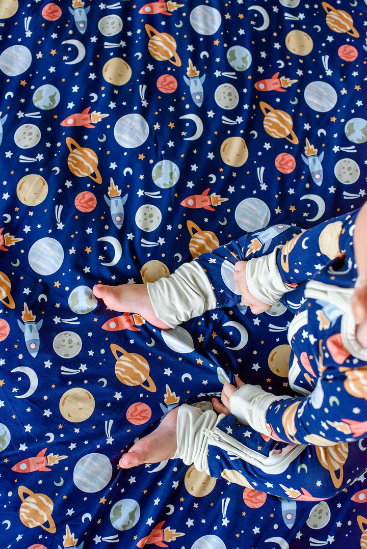 Overhead shot of a baby’s bare feet on a Caden Lane navy knit blanket in a Stellar Snuggles print featuring planets, rockets, and moons, paired with matching pajamas.