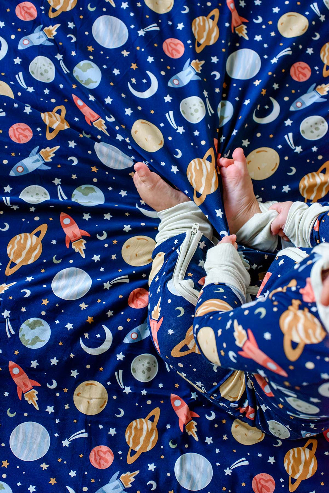 Close-up of a baby gripping a Caden Lane navy knit blanket in a Stellar Snuggles print featuring Saturn, rockets, and shooting stars, paired with matching pajamas.