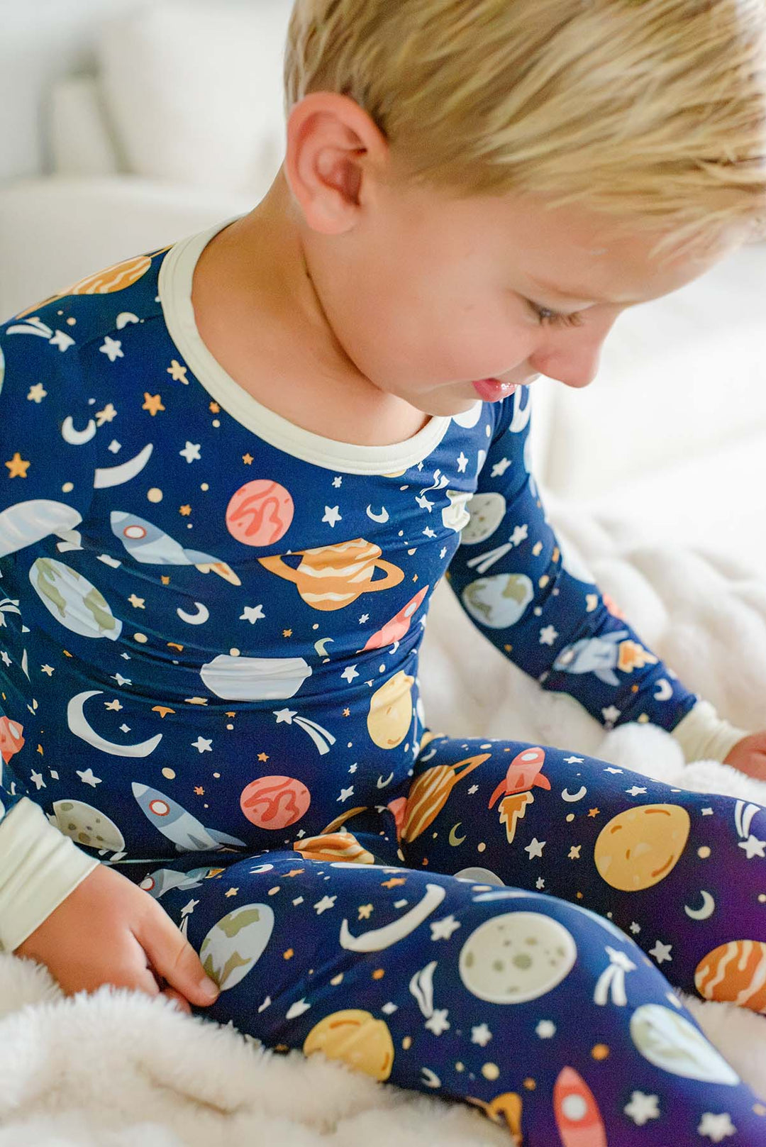 Blond toddler sitting on a fluffy white blanket and looking down, wearing a Caden Lane navy bamboo pajama set in a Stellar Snuggles print featuring planets and rockets.