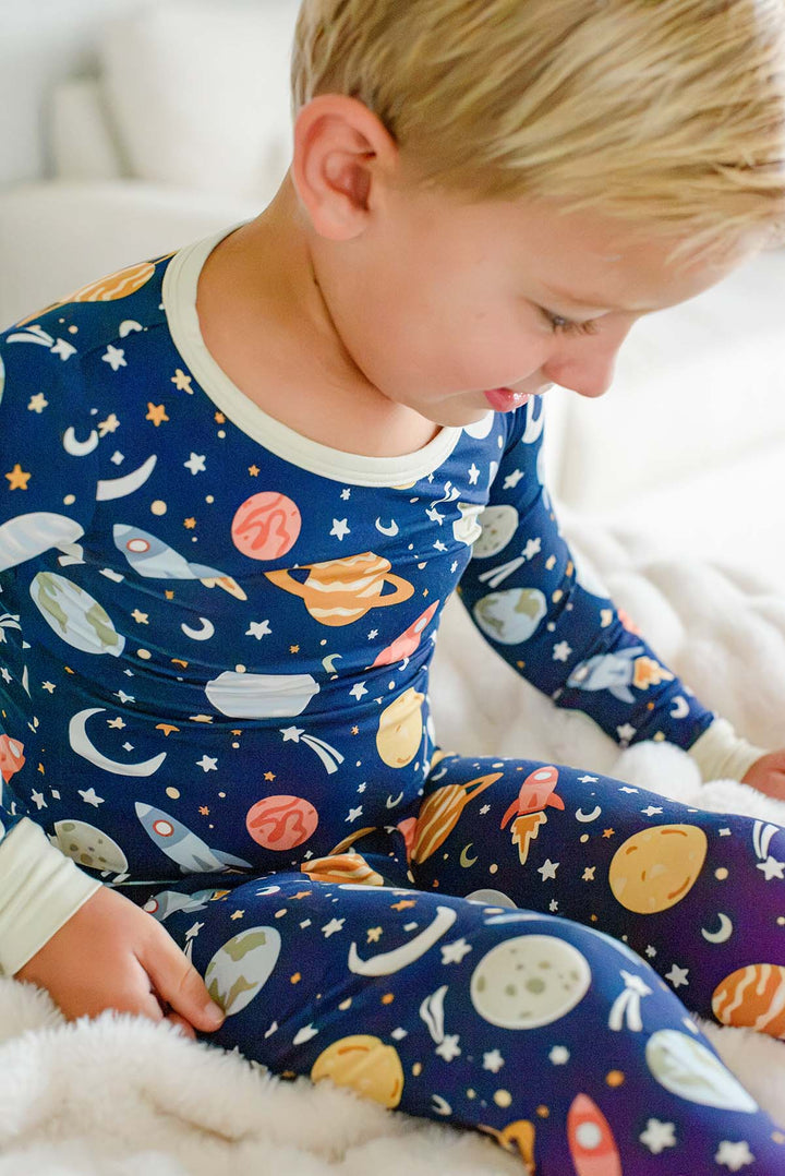 Blond toddler sitting on a fluffy white blanket and looking down, wearing a Caden Lane navy bamboo pajama set in a Stellar Snuggles print featuring planets and rockets.