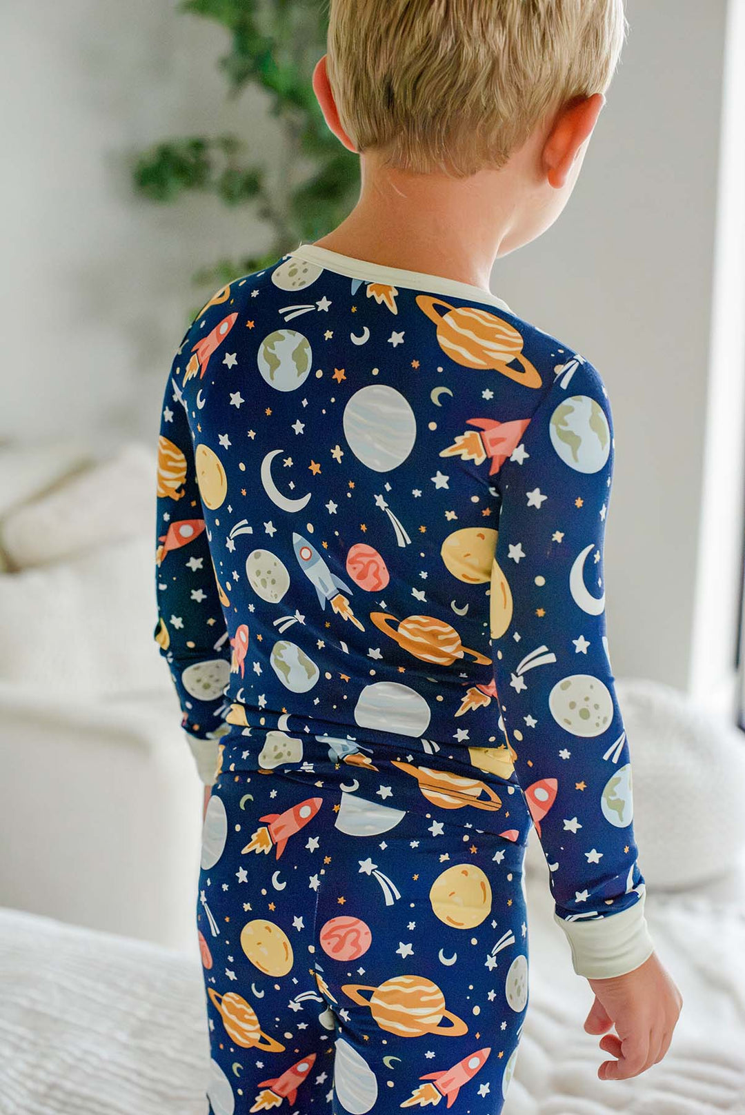 Back view of a blond toddler looking toward a houseplant, wearing a Caden Lane navy bamboo two-piece pajama set in a Stellar Snuggles print featuring rockets, moons, and Earth.